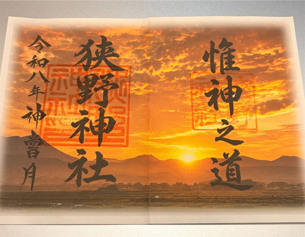 夕日と山並みの写真を背景に「狭野神社」「惟神之道」と書かれた見開きの御朱印