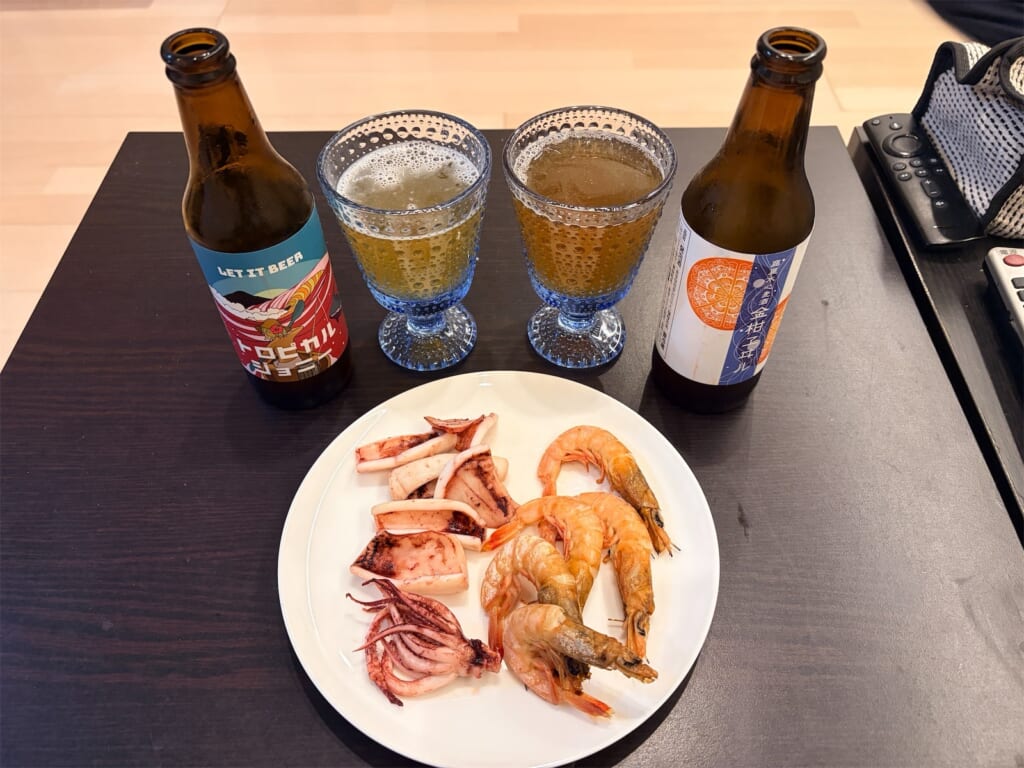テーブルに並んだクラフトビール2本、グラス2つ、焼いたイカとたかえびの皿